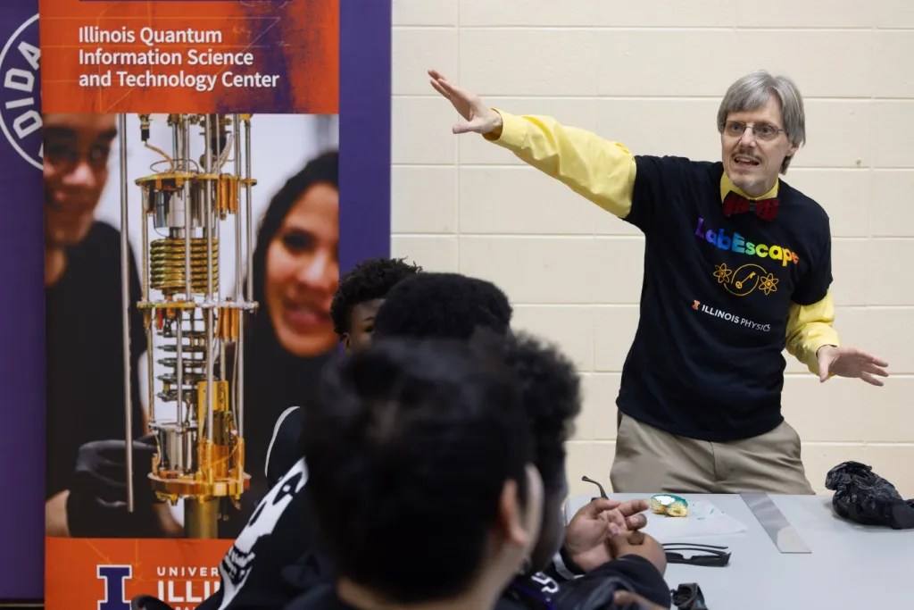 man with grey hair wearing a black t-shirt and bow tie over a long sleeve yellow button down shirt, gesturing at a science poster