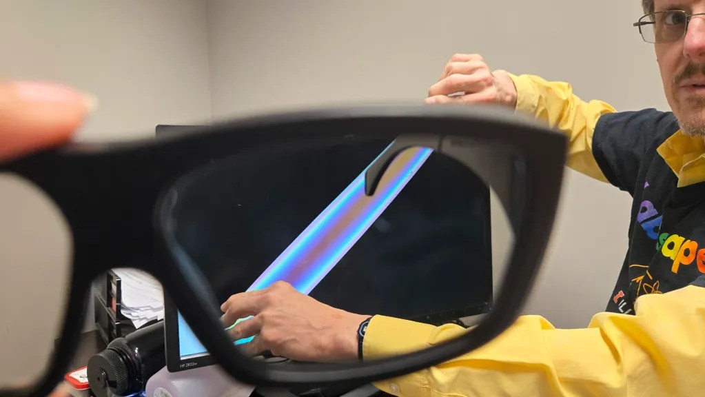 man with glasses wearing a black t-shirt over a yellow button down shirt. He is holding a piece of material in front of a computer screen. When viewed through glasses it shows a prism of color
