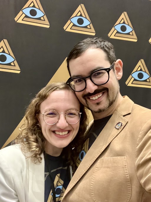 woman with blonde curly hair and glasses, smiling, in a cream color coat. next to her is a man with short brown hair and goatee, wearing glasses, smiling. He is wearing a tan color blazer. Background is an image with logos of a triangle with an eye inside.