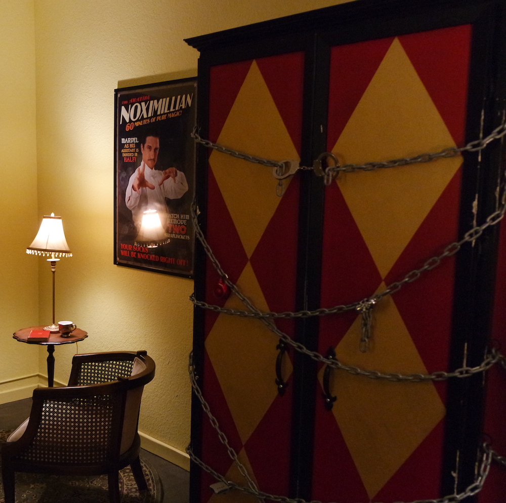 Interior of the Vanishing Act. Depicts massive a red and yellow box chained and locked shut with multiple locks. A poster of Noximillian hangs on the wall beside the box.