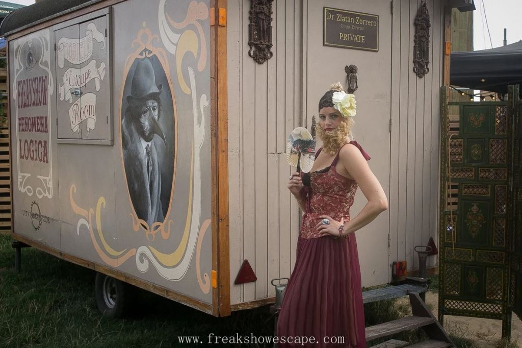Woman dressed as a bearded lady in a wine color corset dress holding a fan, standing in front of a trailer that says "freakshow" on it