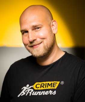 man with shaved head and blond beard smiling against a yellow background. he is wearing a black t-shirt that says "crime runners"
