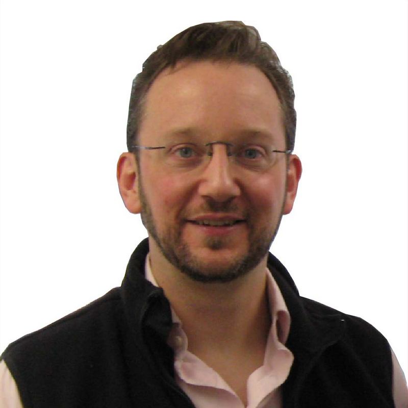 Smiling man with short brown hair and frameless glasses. He has a short beard, and he's wearing a pink button down shirt with a black fleece vest.