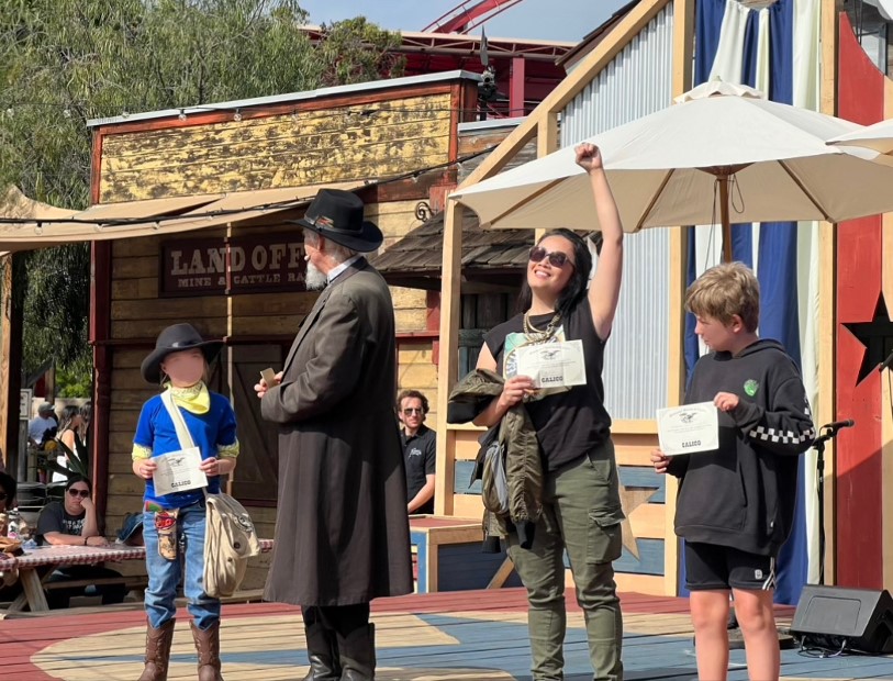 an asian woman on stage with her fist triumphantly in the air. Next to her are bewildered looking children and an older man with a cowboy hat and black duster. they are holding certificates that say "mayor of calico"