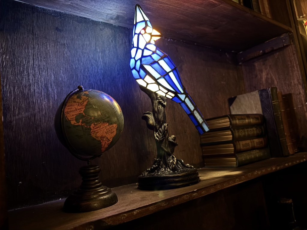 A bird-shaped stained glass lamp on a bookshelf beside a globe and a stack of books.