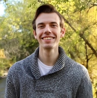 man with styled brown hair, wearing a cowl neck sweater outdoors, smiling.