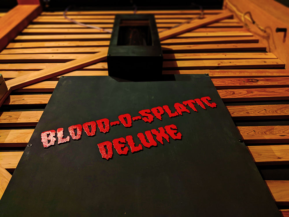 black sign with red words that say "blood o splatic deluxe" on wooden slats