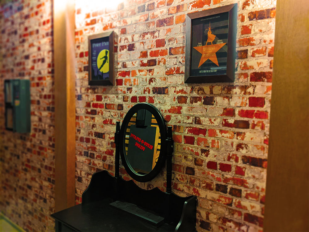 brick wall with posters that resemble broadway posters for shows like peter pan and hamilton. There is also a desk with a mirror.