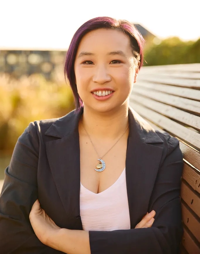 Asian woman with purple hair, smiling, outdoors, wearing a black blazer, white blouse and a necklace with a moon and sun design.