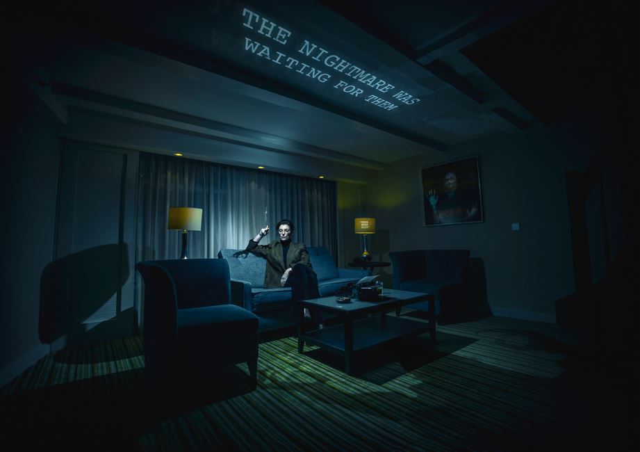 woman in tweed coat sitting in a dark hotel living room smoking. There is the shadow of a monster creeping up on her. Above her is a projection with the words "the nightmare was waiting for them"