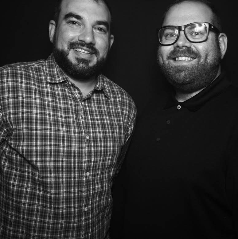 black and white photo of 2 men, both smiling. on the left is a man with a medium length beard, dark hair and wearing a plaid shirt. On the right is a man wearing glasses, a short beard and a dark polo shirt