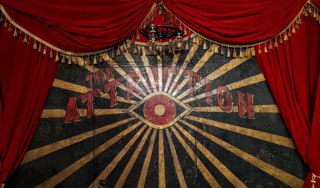 A wall painted with a red, black, and gold eye, bold letters read, "The Attraction." The painting is framed by red and gold curtains.