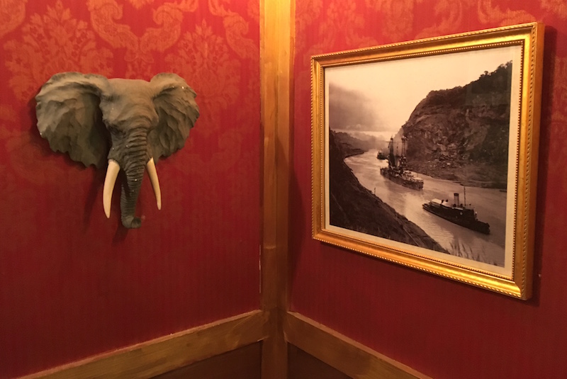 In-game image. A corner with red wallpaper and gold trim. A photo of the Panama Canal hangs on the wall beside an elephant's head.