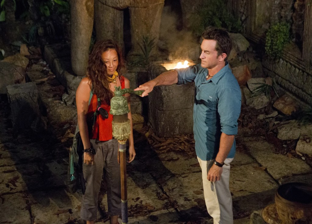 asian woman with wavy hair in a red tank top and grey pants being voted out of Survivor. There is a man in a blue button down safari shirt and khaki pants with brown hair snuffing a torch. They are in a abandoned temple in a jungle setting