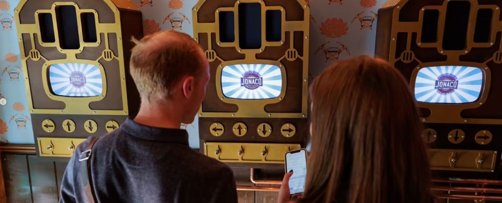 3 steampunk looking boxes with screens that say "jonaco". we see the backs of 2 people standing in front of it while holding a smartphone.