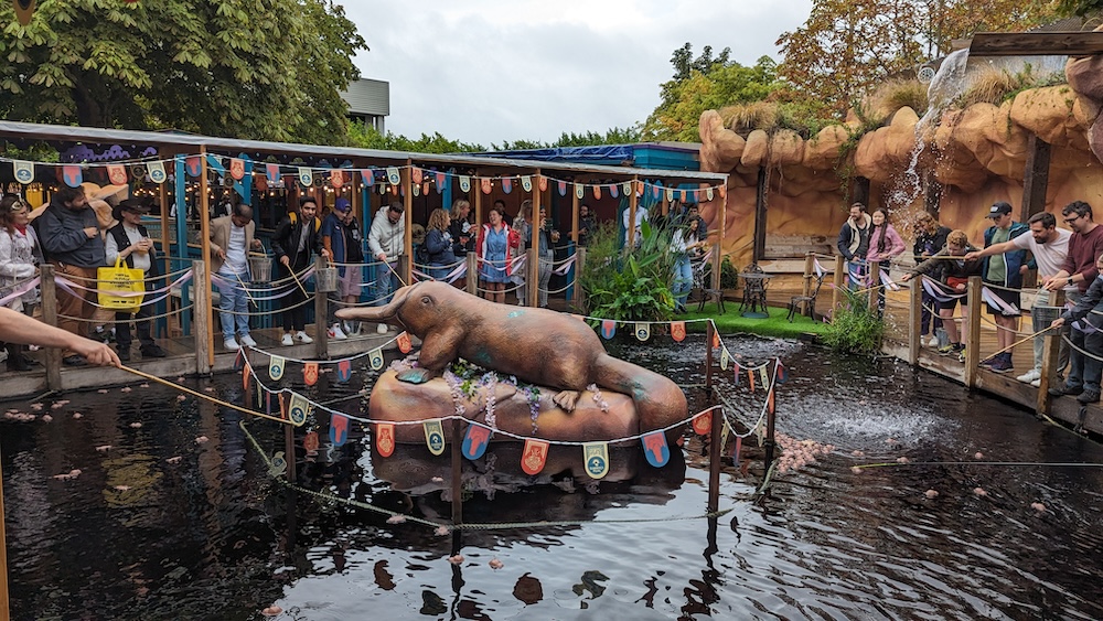 A pond with a large platypus statue in the middle. On the banks of the pond are many people fishing for small platypuses.