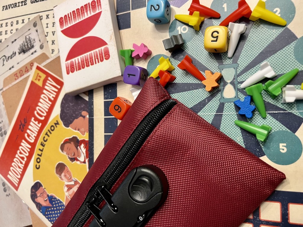Close up of game conponents. A maroon bag with a lock, some meeple, rock game pieces, a game board, and a magazine for "The Morrison Game Company Collection"
