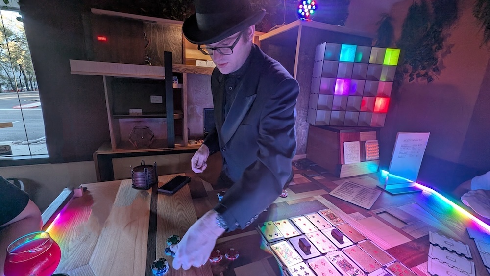 A man in a black suit and hat at a wooden bar, manipulating poker chips beside a card game and puzzle boxes.