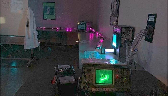 room with stainless steel tables. There are scientific instruments on the tables, and a whiteboard in the room with a labcoat hanging from it.