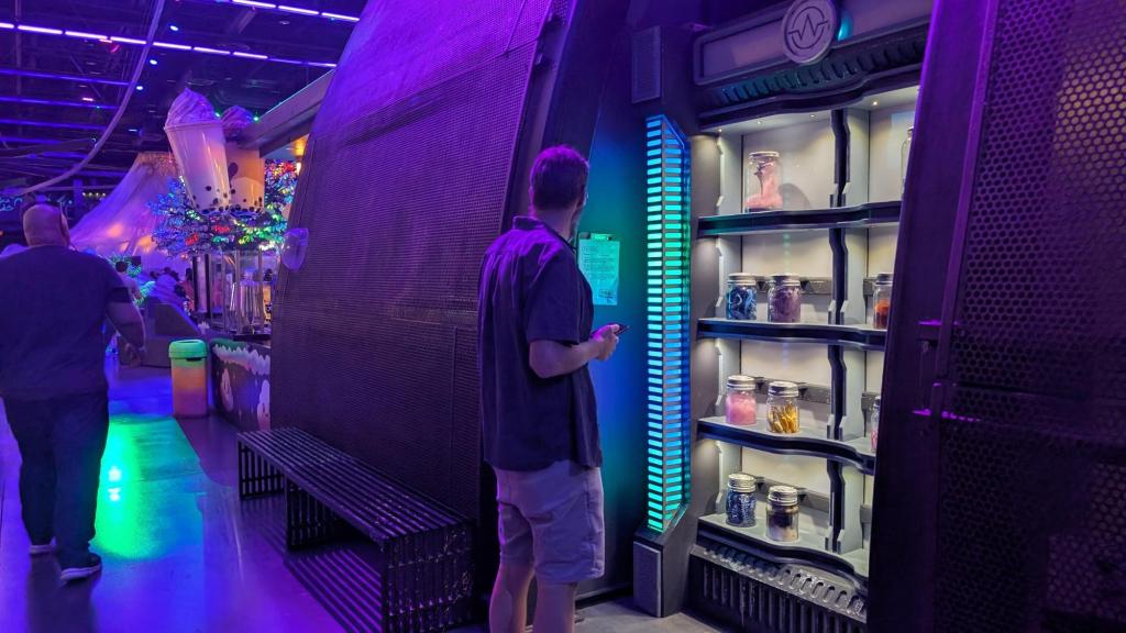 A man stands in front of a lighted shelf holding jars of colorful alien objects and reads a clipboard mounted on the wall beside it. It is in an open area of a large entertainment district