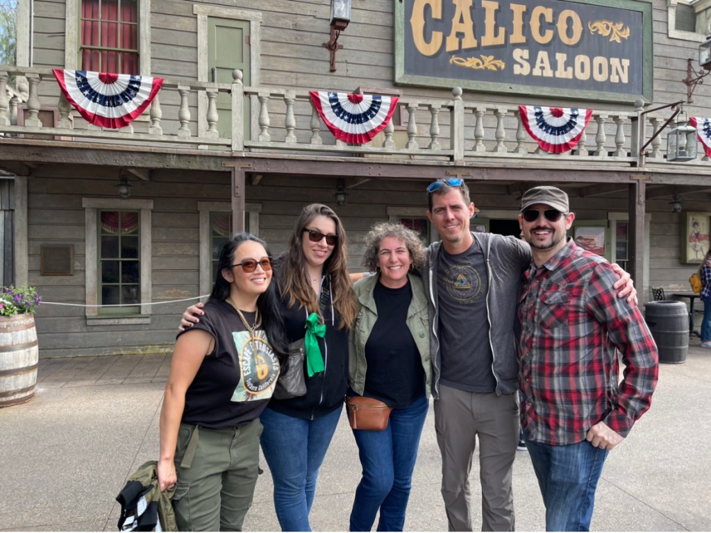 Group of friends smiling in front of an old western style saloon. From left to right is an asian woman in black t-shirt, green cargo pants, a woman with jeans and a green bow on her bag with long brown hair. a woman with a green jacket, black shirt and jeans with shoulder length curly hair, a tall man in grey t-shirt and hoodie with grey pants, and a man with a plaid shirt, hat, goatee and jeans.