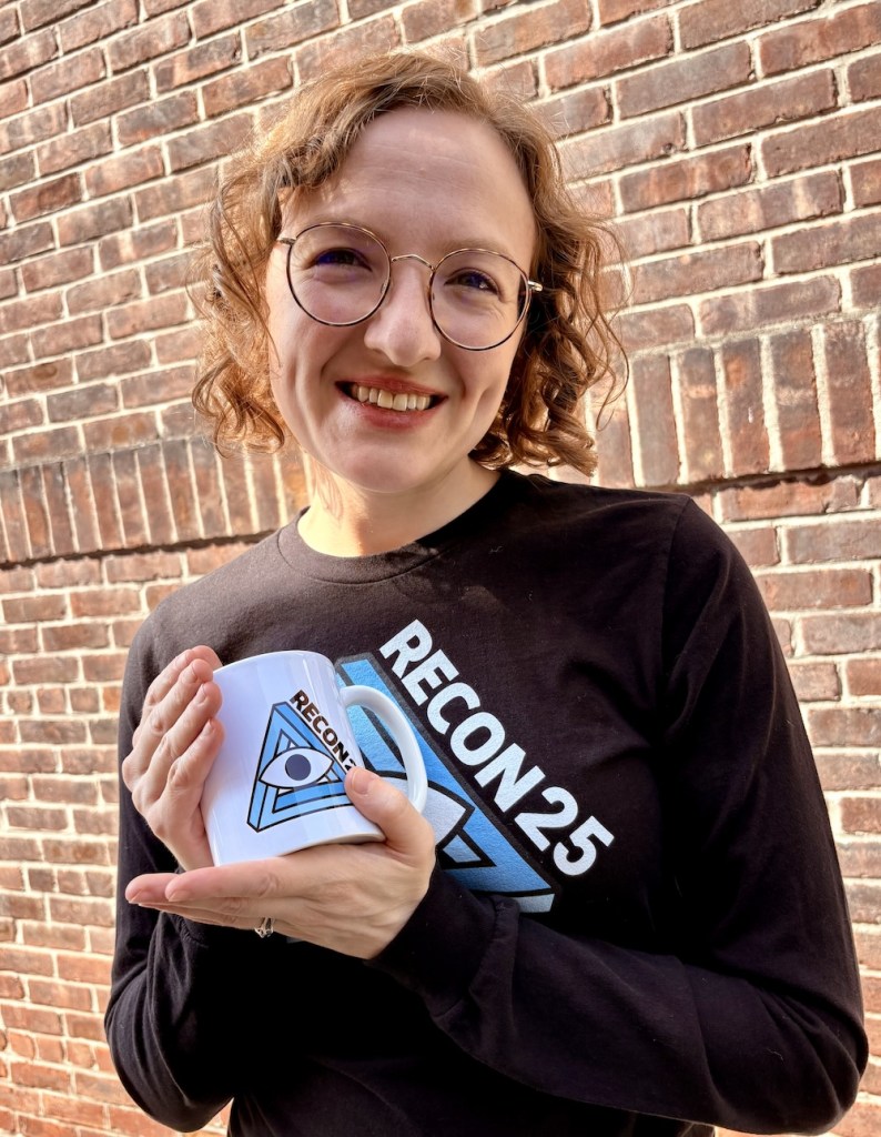 Blonde woman in glasses wearing a black RECON 25 shirt and holding a white RECON 25 mug.