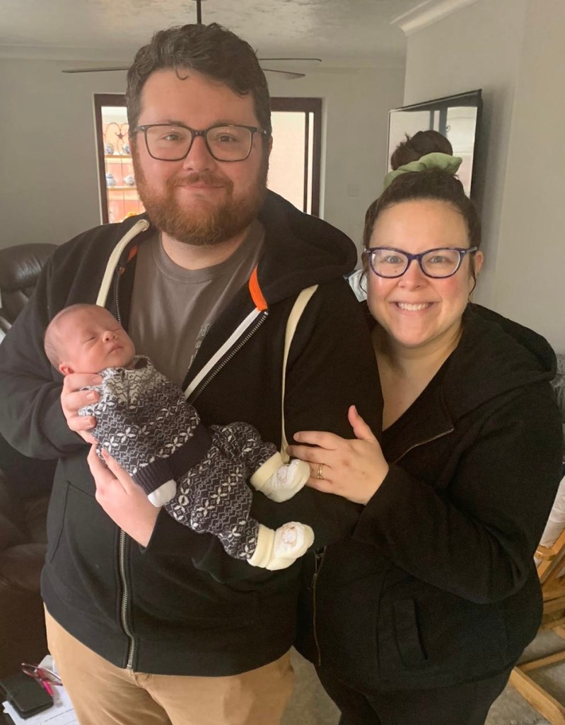 family portrait photo of a man with short brown wavy hair and a short beard in glasses. He is wearing a black hooded sweatshirt, grey shirt and khaki pants. He is holding a newborn infant wearing a sweater set. There is a woman with her brown hair in a bun, smiling, in glasses. She is wearing a black hooded sweatshirt and holding onto the man's arm.