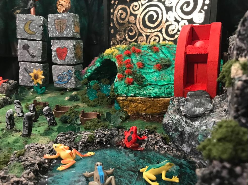 image of a diorama from the online game "keeper and the fungus among us". it's a woodland scene with a red watermill next to a green cave. in the front there is a fake miniature pond with colorful toy frogs. there are also buckets set into the ground, some stone blocks with symbols on them stacked vertically in the back.