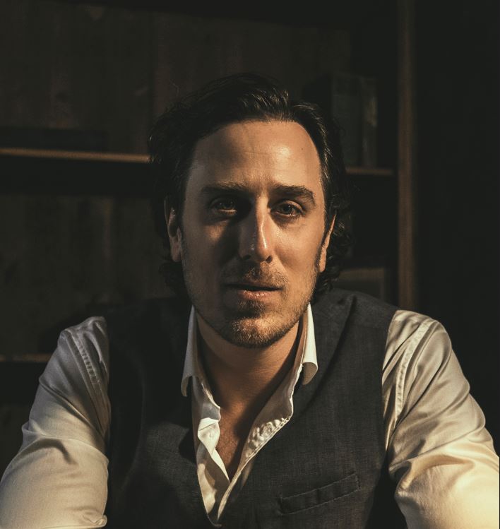 slender man sitting at a desk with hair combed back from his face, and an intriguing expression. He is wearing a white button down shirt with a grey vest. He has a short beard, and he is dramatically lit with dark bookshelves behind him.