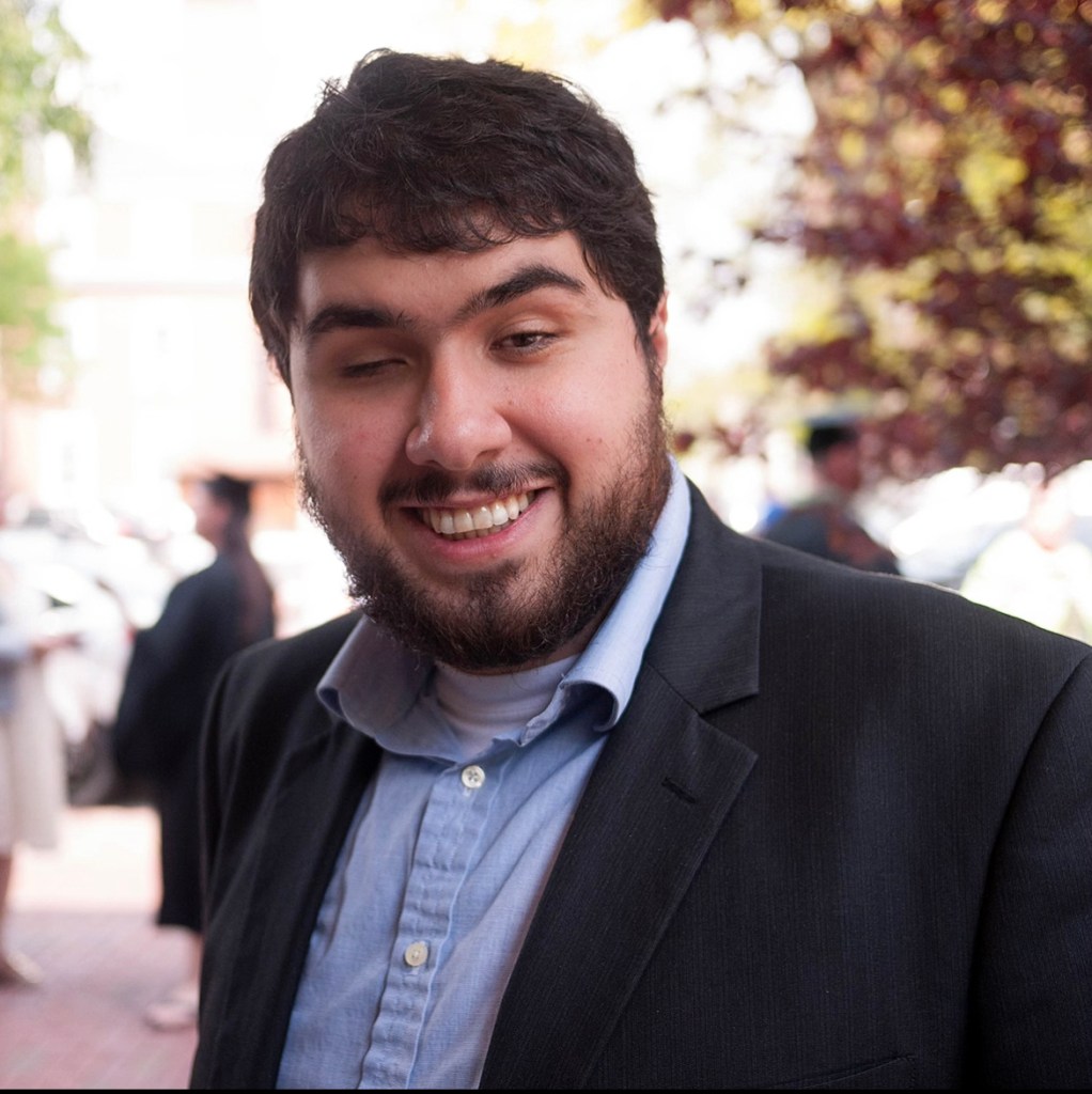 The headshot of a light skinned Persian man with a toothy grin. He has a full beard, thick wavy dark brown hair and his right eye is closed. He wears a charcoal grey sports coat over a light blue button down shirt.