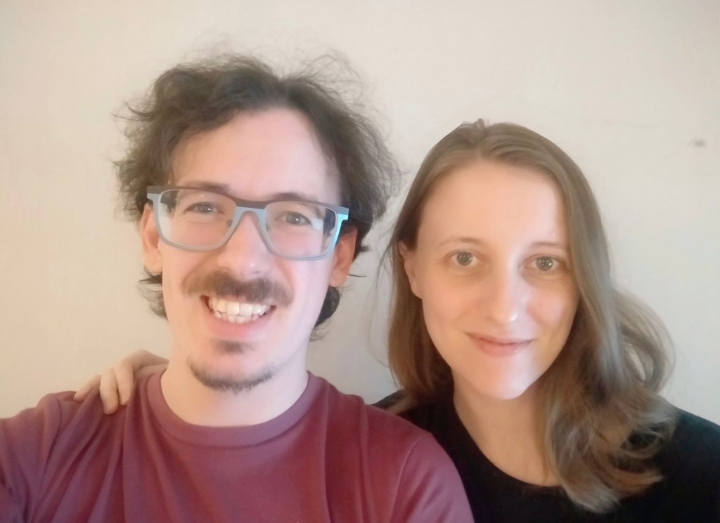 australian man and woman smiling. The man has wavy brown hair, glasses, and a roguish grin. He has a short goatee and is wearing a maroon t-shirt. The woman has dark blonde hair and blue eyes. She is smiling and wearing a black shirt.