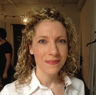 Smiling woman with curly shoulder length blond hair and grey eyes. She is wearing a white button down shirt.