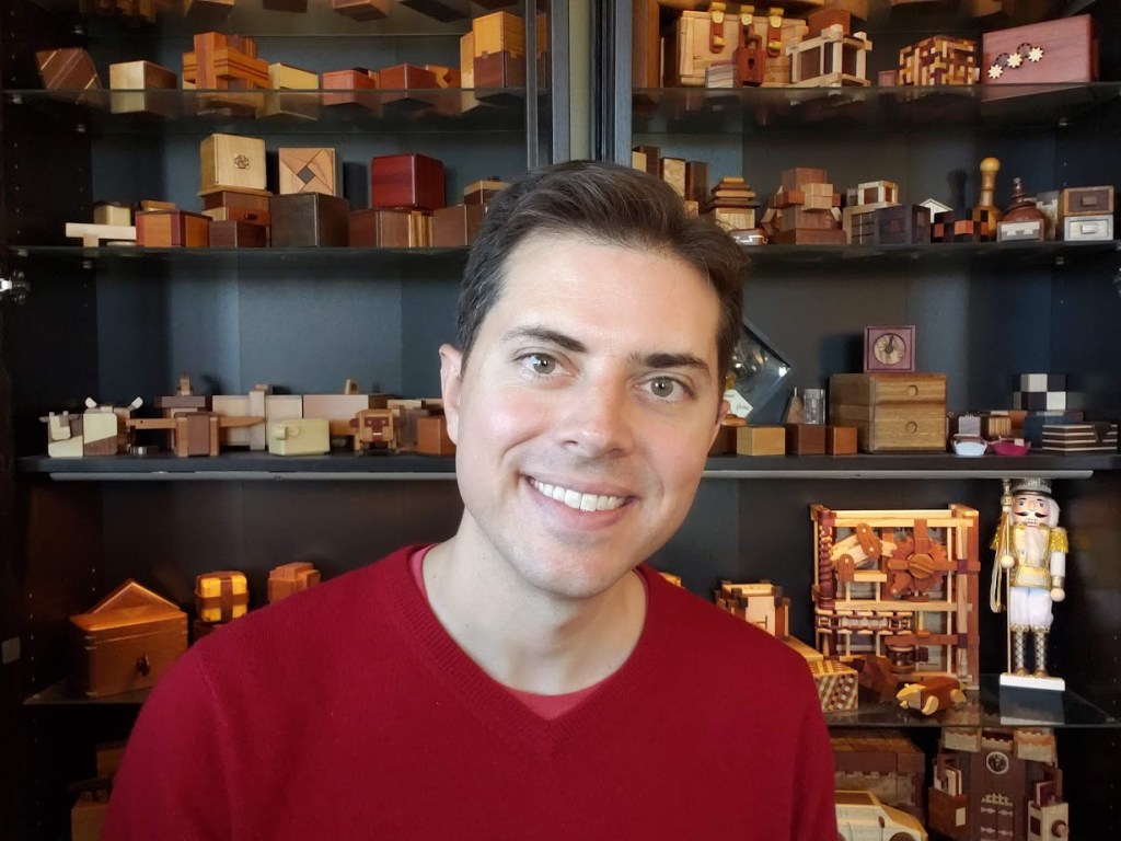 smiling man with neat brown hair and a red sweater in front of shelves filled with wooden mechanical puzzles.