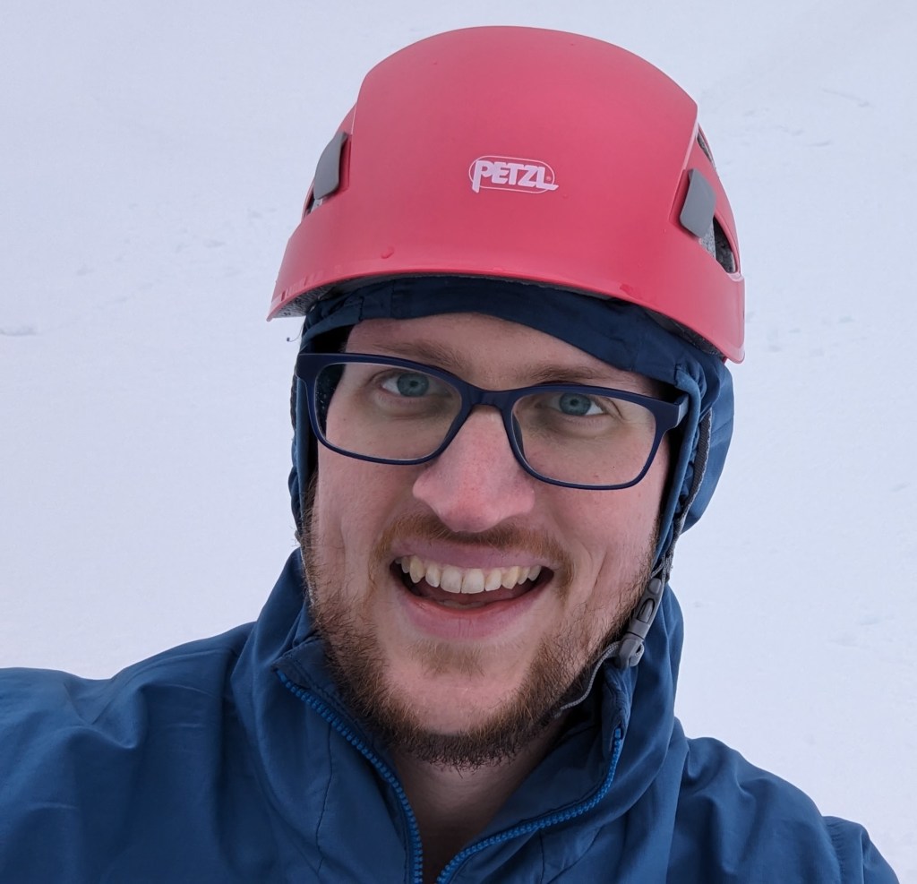 smiling man with a brown beard wearing glasses, blue hooded jacket and a pink climbing helmet