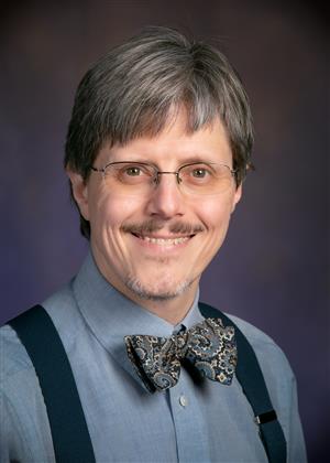 man with grey hair wearing glasses, blue shirt, paisley bowtie, blue suspenders. He has a goatee and is smiling.