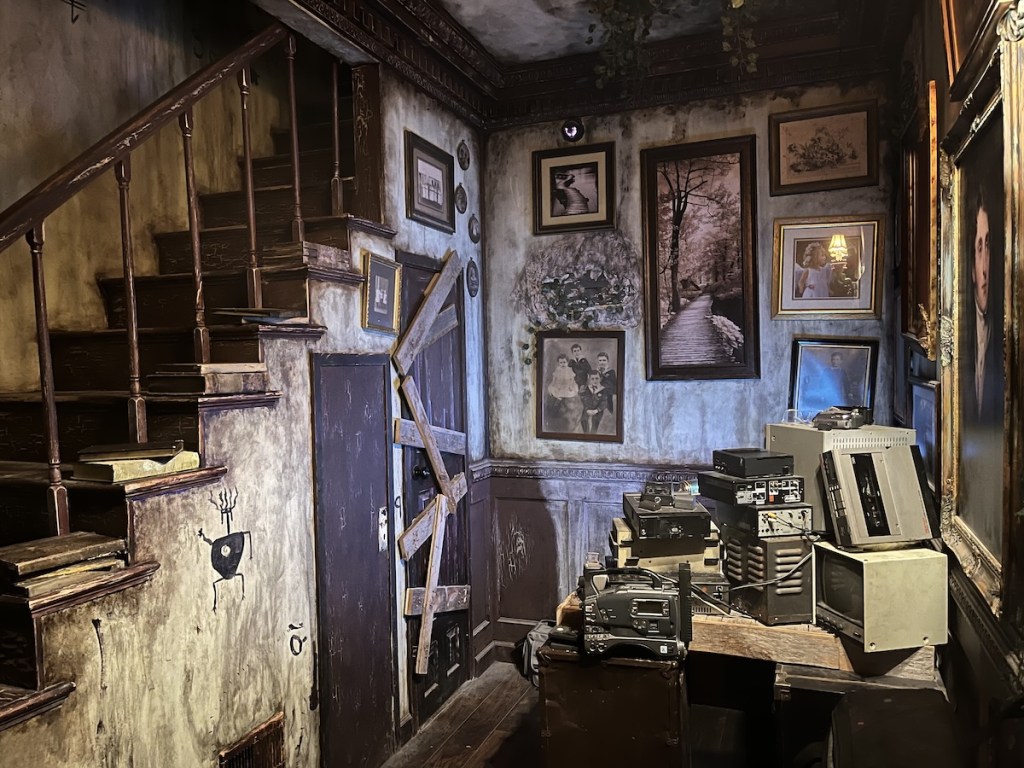 An old room with framed paintings on the walls, a desk covered in technology, and stairs leading up.