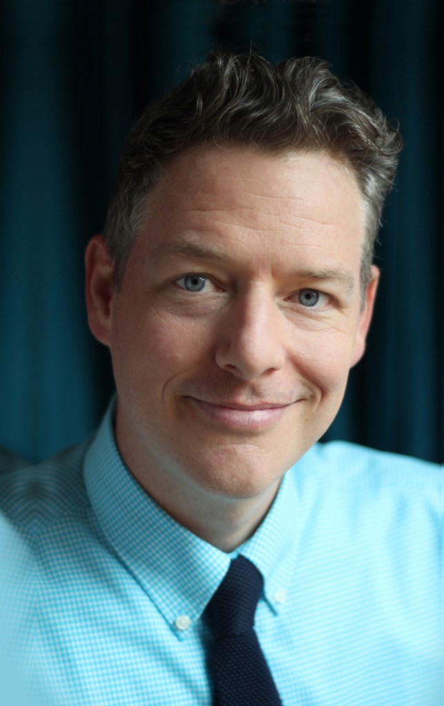 caucasian man smiling. he has neat wavy brown hair, blue eyes and is clean shaven. He is wearing a blue checked button down shirt with a black tie.
