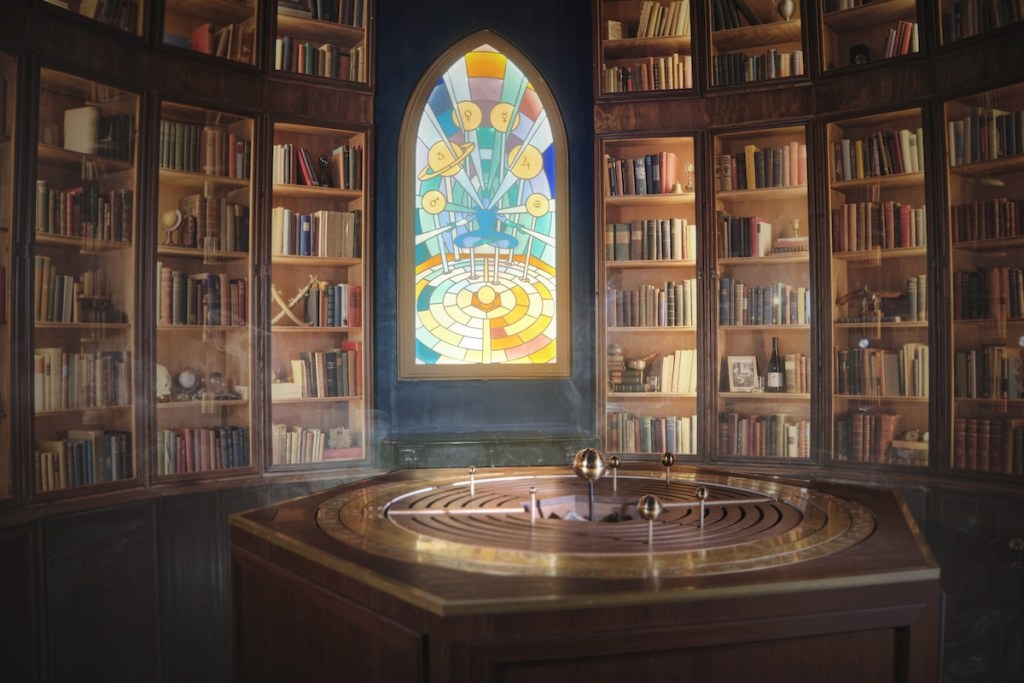 A curved wall of bookshelves that goes up to the ceiling, with a stained glass window in the middle and an orrery in front.