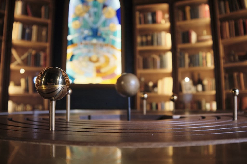 The image is focused on the orrery in the foreground. Blurred in the background is a wall of bookshelves with a stained glass window in the center.