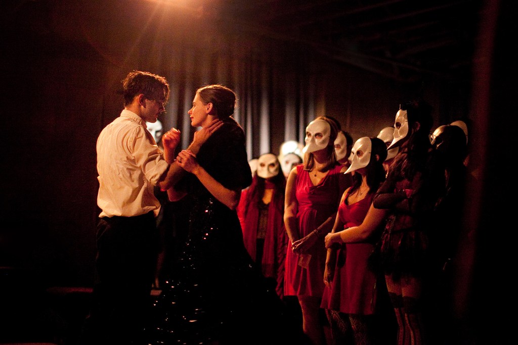 man and woman facing each other, he has his hand on her throat. she has her brunette hair up and is wearing a sequined evening gown. He is in a white button down shirt with the sleeves rolled up and black pants. There is an audience surrounding them, wearing red dressed and white masks.