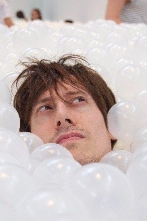 man with brown hair with only his face showing. He us surrounded by translucent white balls that give the appearance of bubbles