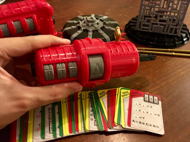 Quiz Master: A red tube with input for a 3 digit number and a letter answer. A key is protruding from its side. An array of corresponding puzzle cards are fanned in front of it.