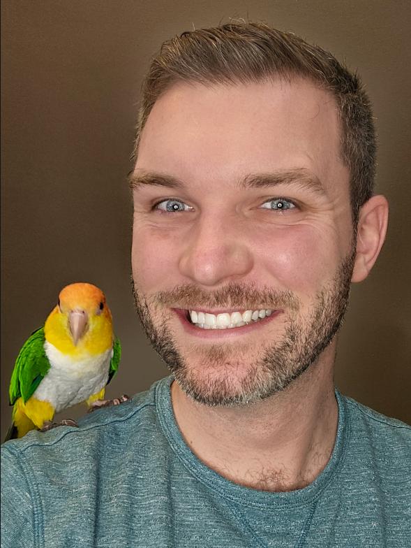 man with blonde hair and short beard, icy blue eyes, wearing grey shirt with a bird on his shoulder