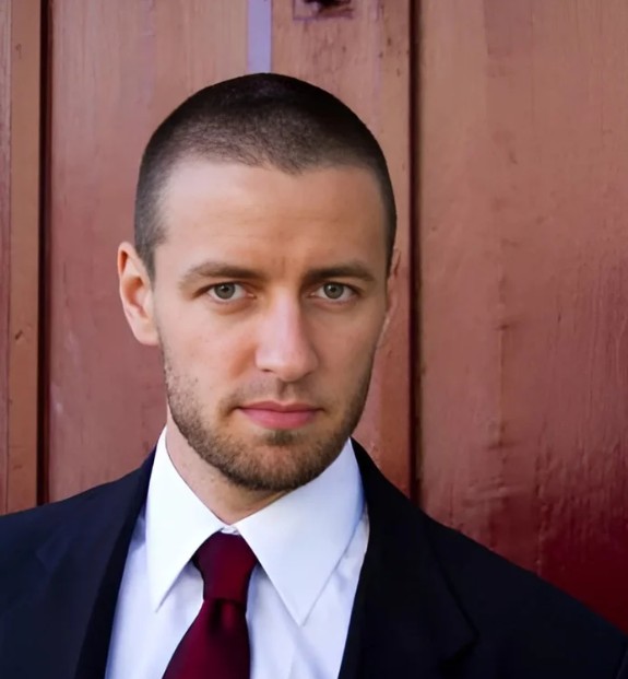 man with very short hair and short half beard in a suit with red tie