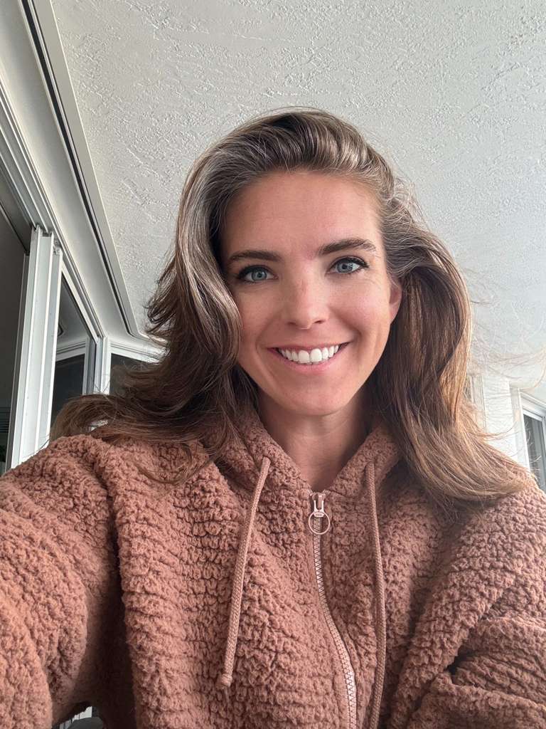 woman with dark blonde hair, blue eyes, smiling, wearing a dusty pink zip up sweatshirt.