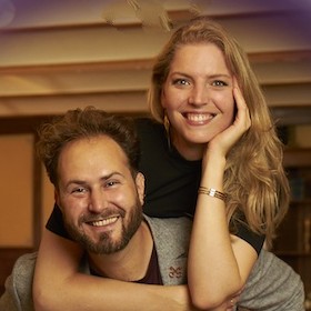 man with light brown hair and beard smiling in a grey blazer with a blond woman smiling in a short sleeve black turtleneck propped over his shoulder
