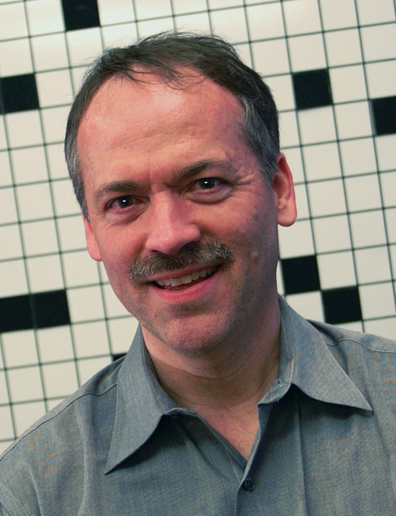 middle aged man with salt and pepper hair, mustache, smiling, wearing a grey button down shirt, in front of empty crossword puzzle grid