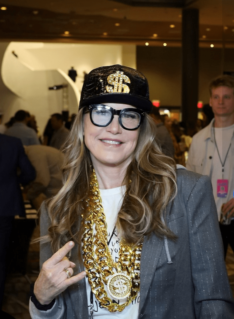 woman with long blond hair with black glasses wearing large gold chains and a black cap with a large gold dollar sign on it.