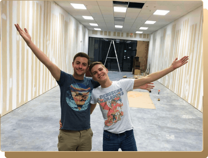 Two young men standing in front of an unfinished, empty room that is still in the process of construction. They have arms around each other's backs with outside arms thrown up and out. On the left, he has neat brown hair parted on the side, smiling, wearing a blue graphic tee and khakis. On the right, he has neat blonde hair, wearing a white t shirt with a Dungeons and Dragons graphic and blue jeans.