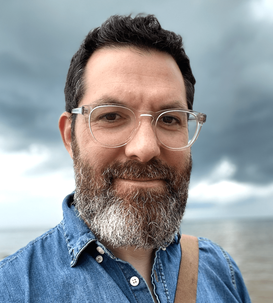 man with short neat dark hai and a salt and pepper beard wearing clear plastic glasses and a denim shirt